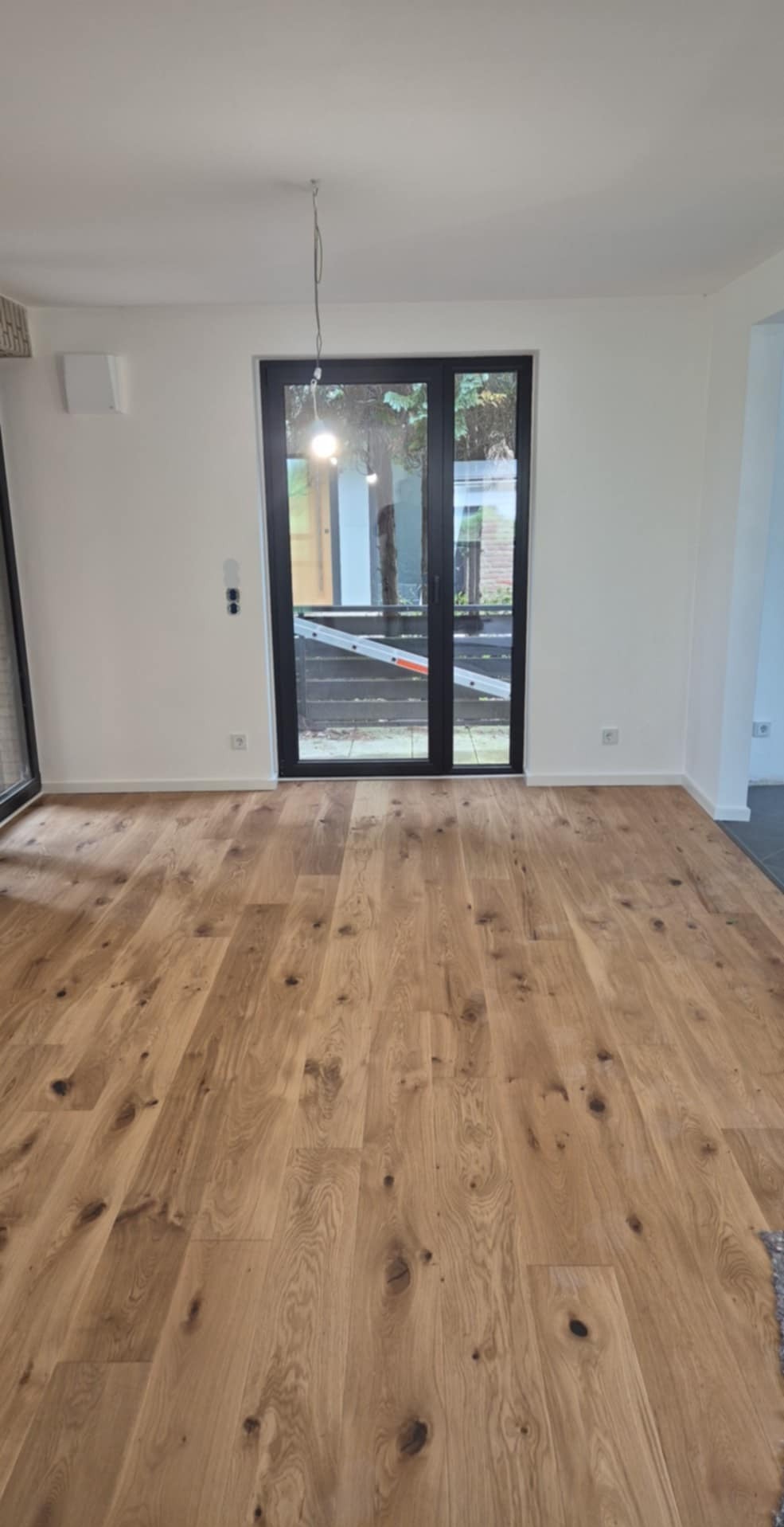 Jasny parkiet w nowym wnętrzu z czarnymi ramami okiennymi i drzwiami balkonowymi. Widoczna drabina na zewnątrz. Minimalistyczny design.