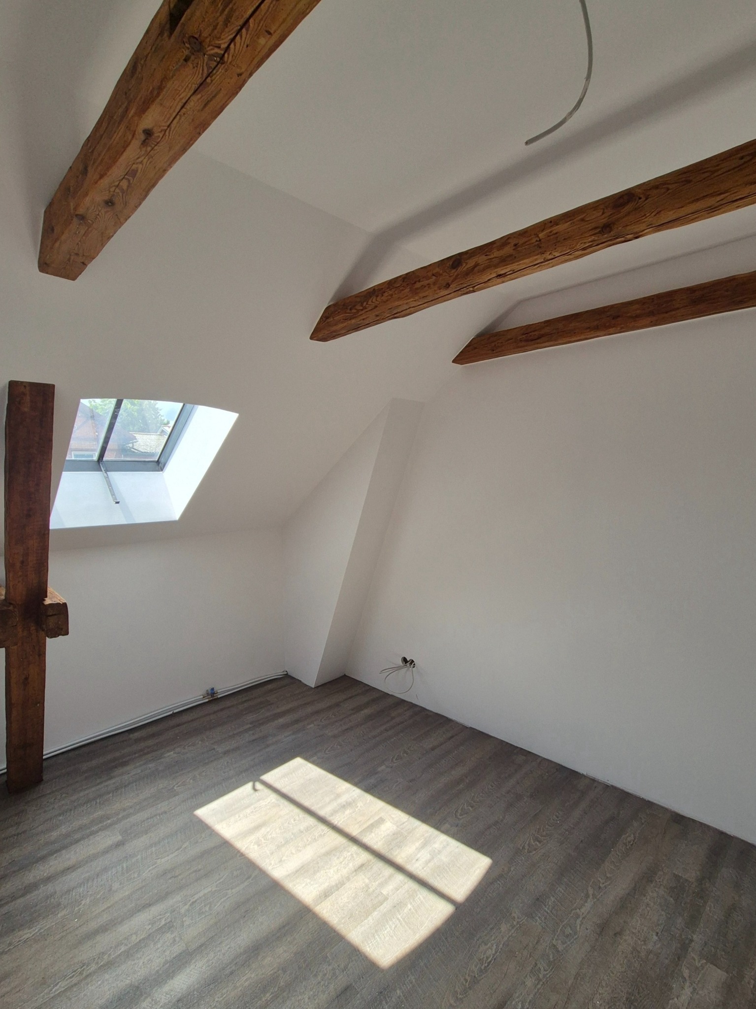 Jasne poddasze z drewnianymi belkami stropowymi, oknem dachowym i panelami podłogowymi w odcieniach szarości. Widoczne gniazdka elektryczne i fragment instalacji.