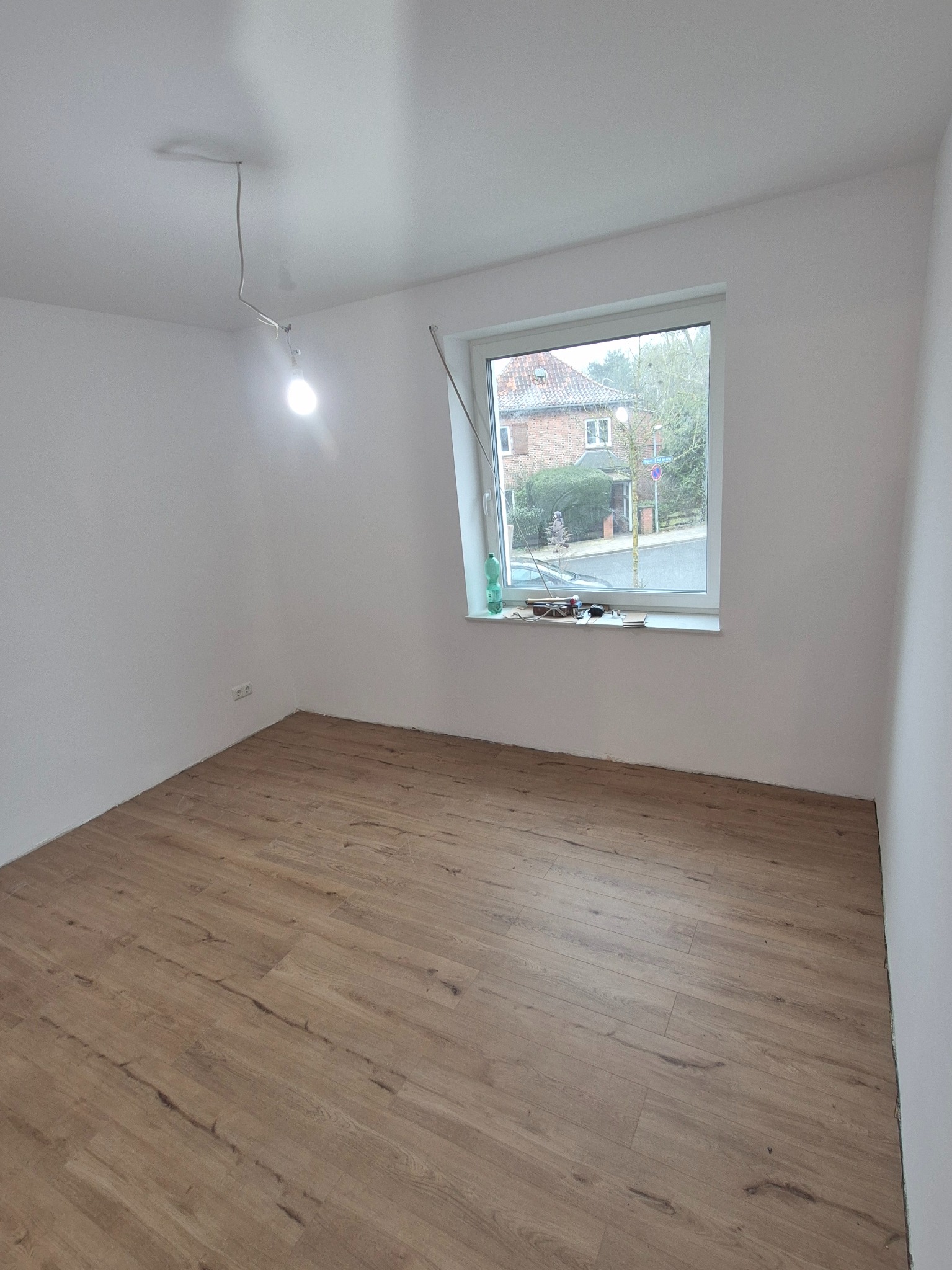 Jasne wnętrze po ułożeniu paneli podłogowych. Widoczne narzędzia na parapecie i niedokończone oświetlenie sufitowe. Świeżo wykończone ściany.