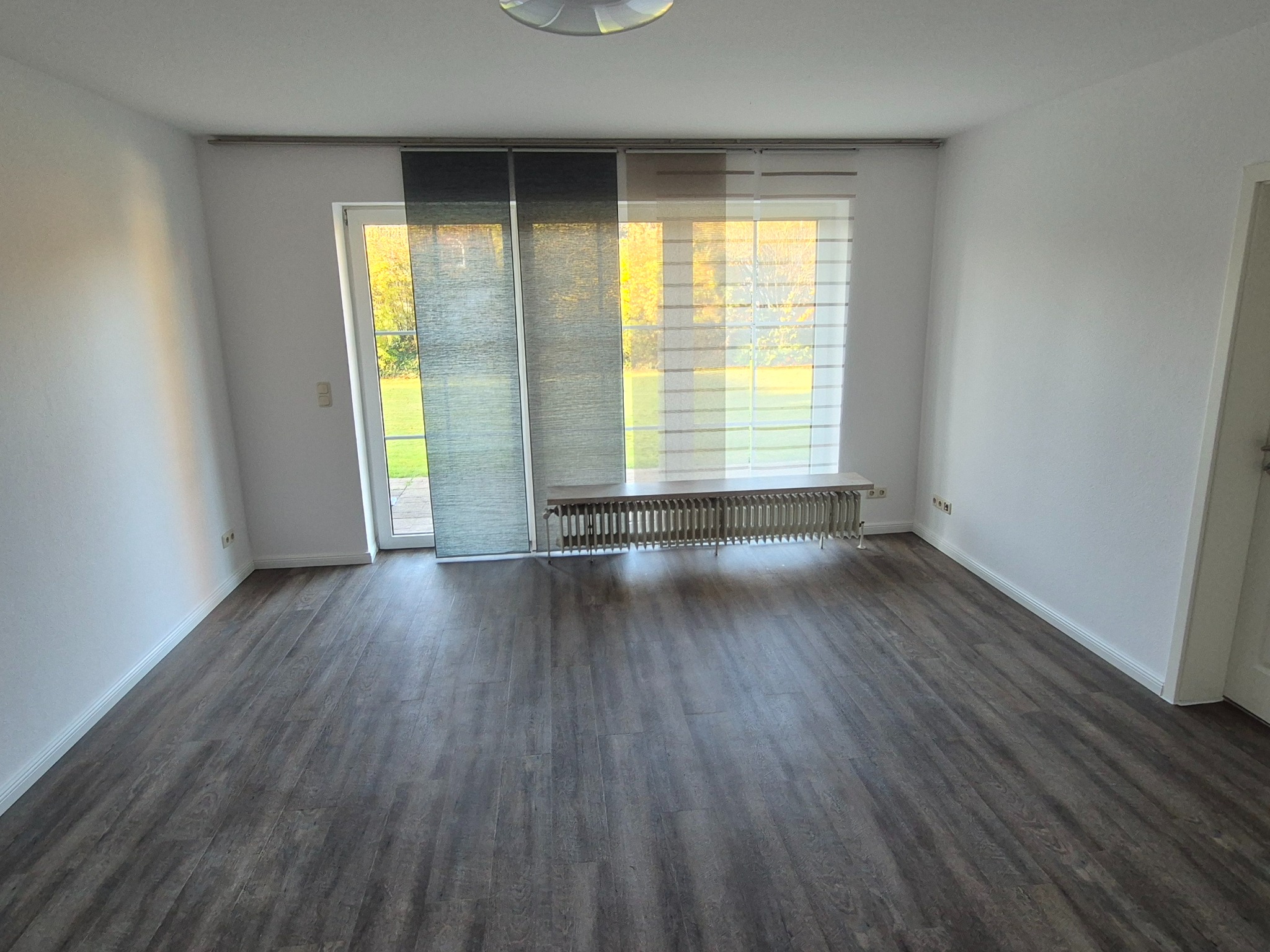 Pokój z panelami podłogowymi w odcieniach szarości, białe ściany, okno z żaluzjami panelowymi i grzejnik. Widok na zewnątrz przez otwarte drzwi balkonowe.