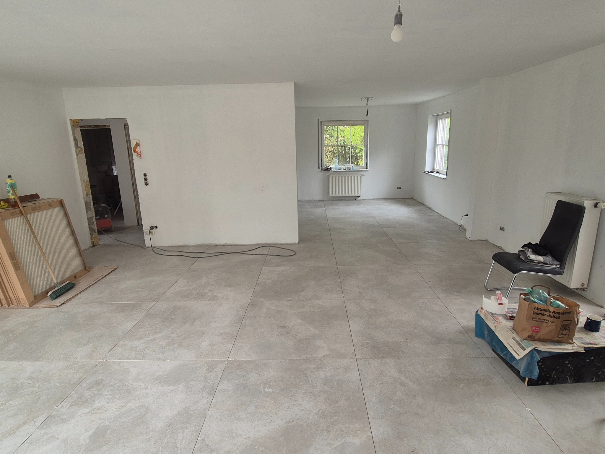 Jasnoszara, duża podłoga z płytek w przestronnym, odświeżonym pokoju. Widoczne przygotowania do dalszych prac wykończeniowych, takie jak malowanie ścian i montaż drzwi.