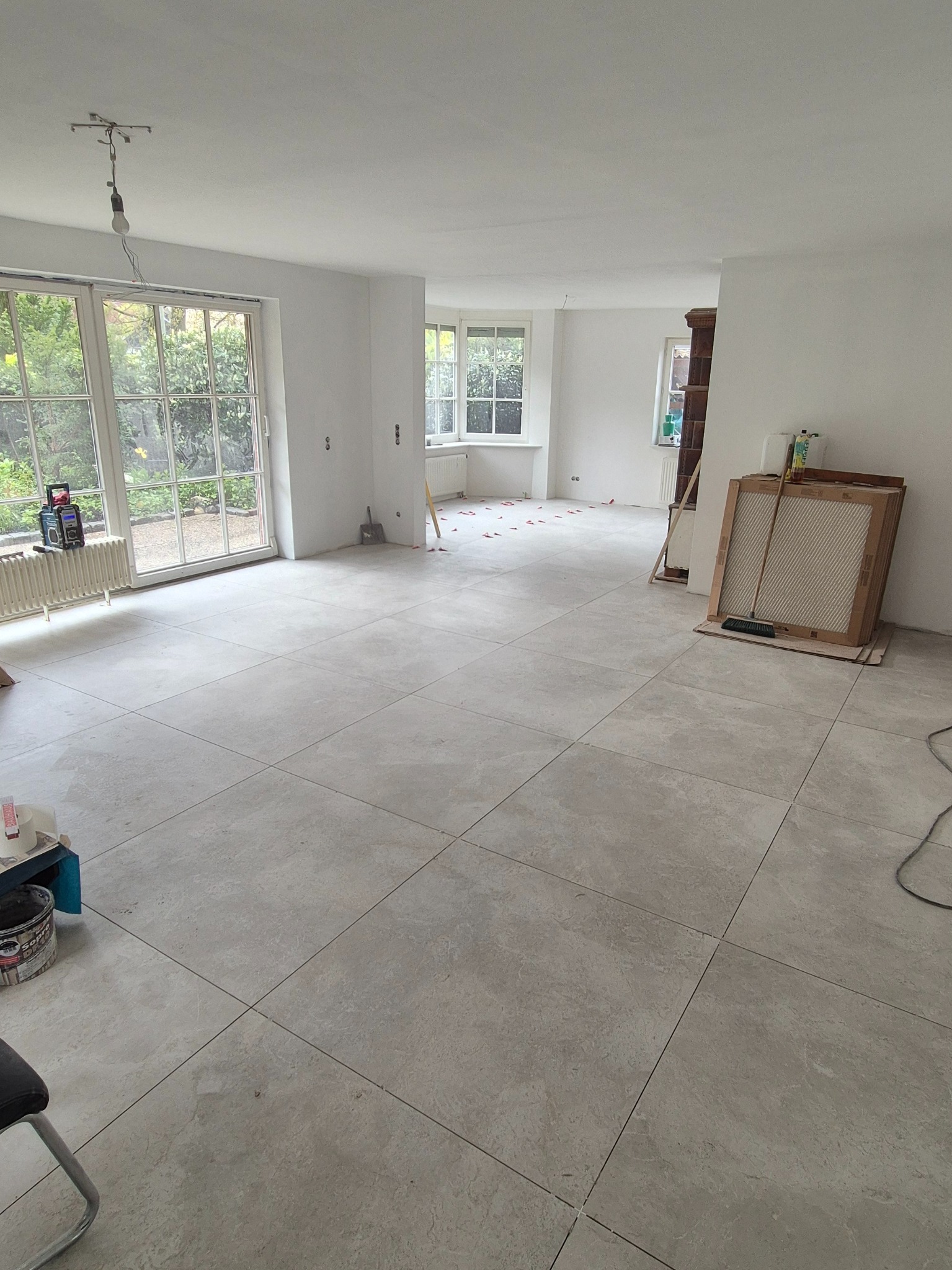 Jasnoszara, duża płytka podłogowa w przestronnym, jasnym pokoju z dużymi oknami. Widoczne narzędzia i materiały budowlane, sugerujące etap prac wykończeniowych.