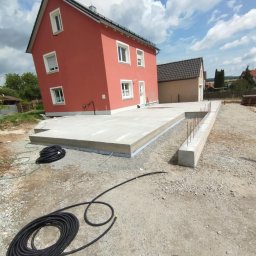 Radbud - Wylana płyta fundamentowa pod dom jednorodzinny z widocznymi zbrojeniami i izolacją, na tle czerwonego budynku w słoneczny dzień. Widoczne węże i narzędzia budowlane.