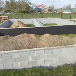 Radbud - Wykopany i częściowo wymurowany fundament pod budynek z bloczków betonowych. Widoczna izolacja pionowa fundamentu. W tle krajobraz wiejski.