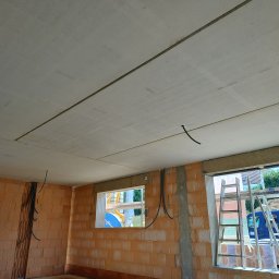 Radbud - Surowy stan wnętrza domu w trakcie budowy: ściany z cegły, betonowy strop z widocznymi przewodami elektrycznymi, drabiny opierające się o okno.