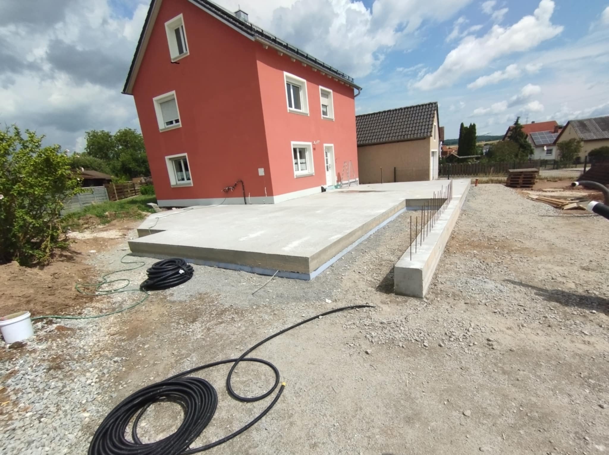 Wylana płyta fundamentowa pod dom jednorodzinny z widocznymi zbrojeniami i izolacją, na tle czerwonego budynku w słoneczny dzień. Widoczne węże i narzędzia budowlane.