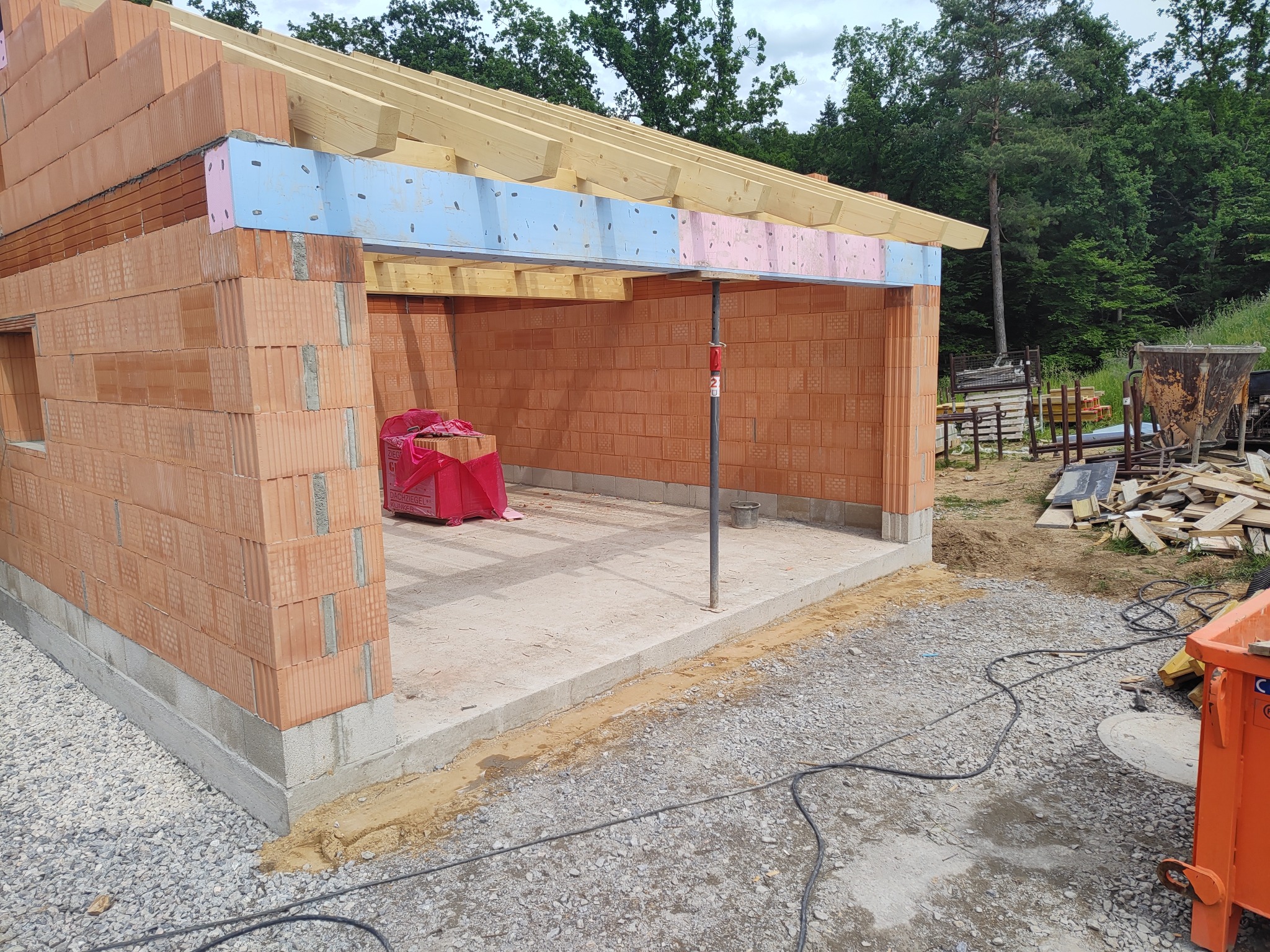 Budowa garażu z czerwonej cegły: ściany w trakcie wznoszenia, widoczne drewniane belki stropowe i izolacja. Na podłożu gruz i materiały budowlane.
