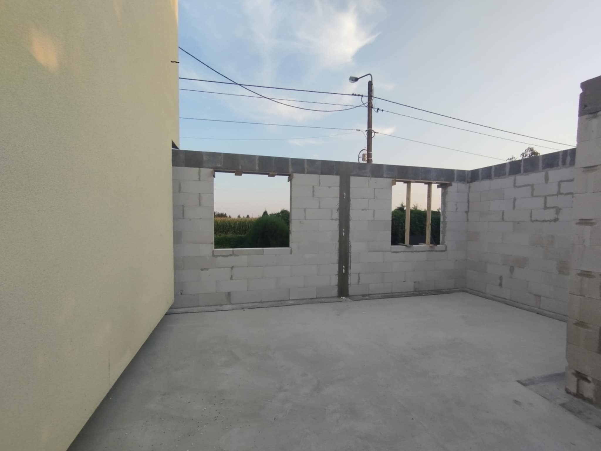 Wnętrze surowego domu w trakcie budowy: ściany z betonu komórkowego z otworami okiennymi, w tle zieleń. Widoczne ślady zaprawy. Stan surowy zamknięty.