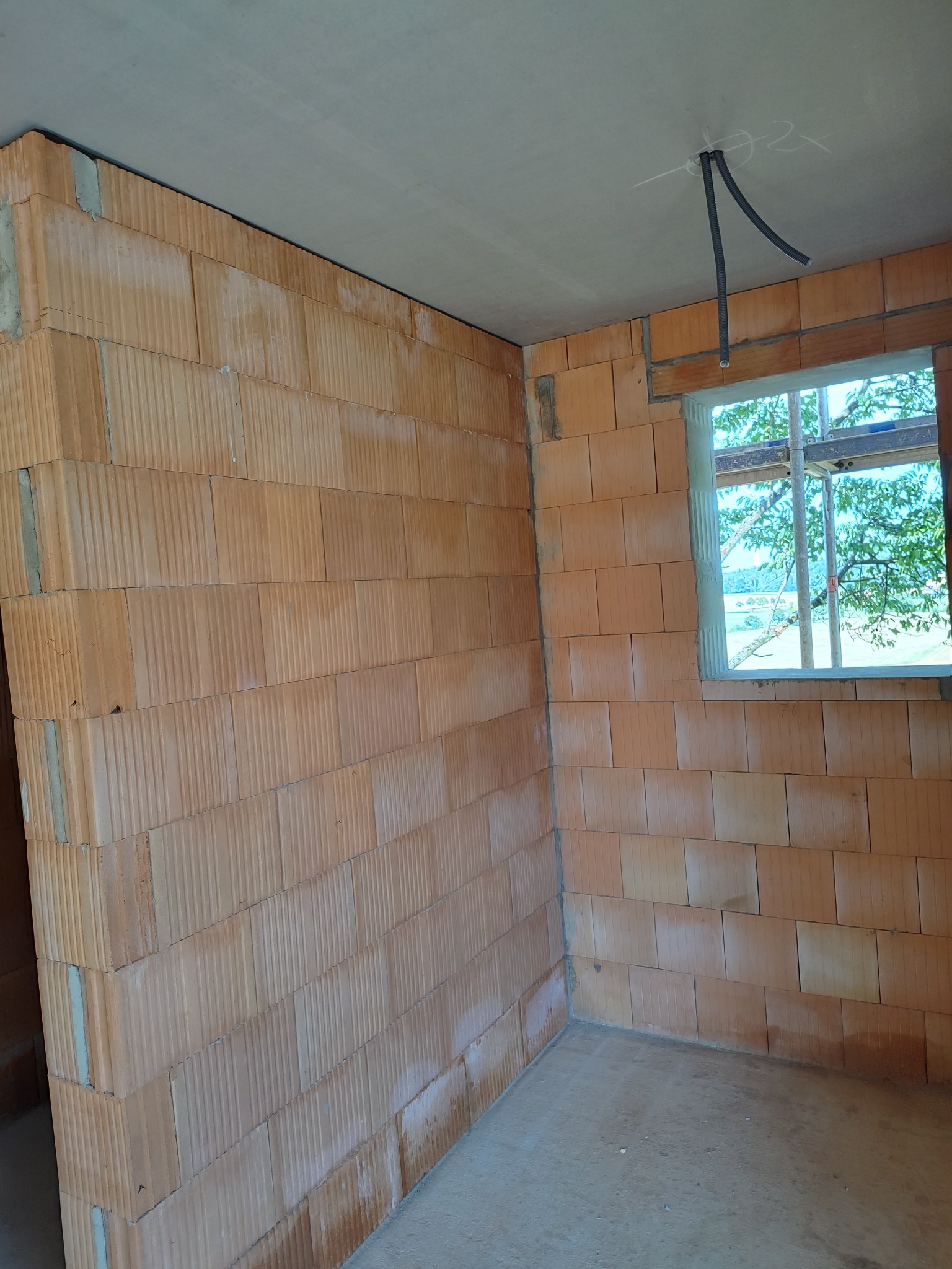 Wnętrze w trakcie budowy: ściany z czerwonej cegły, okno z widokiem na zieleń i jezioro, wystające rury instalacji elektrycznej z sufitu.