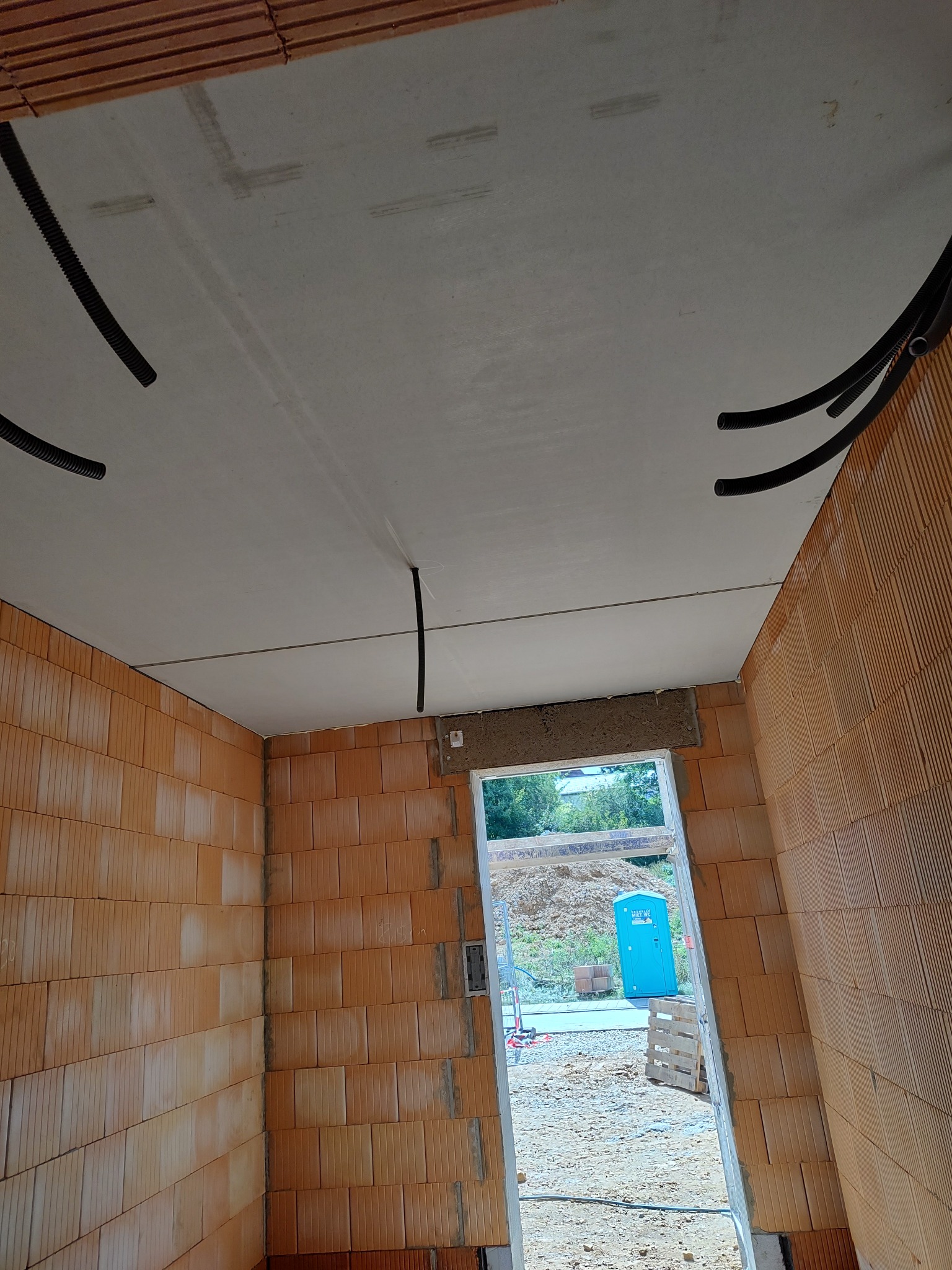 Wnętrze surowego domu z pustaków ceramicznych, widoczne rury instalacyjne pod sufitem i otwór drzwiowy z widokiem na zewnątrz. Etap budowy.