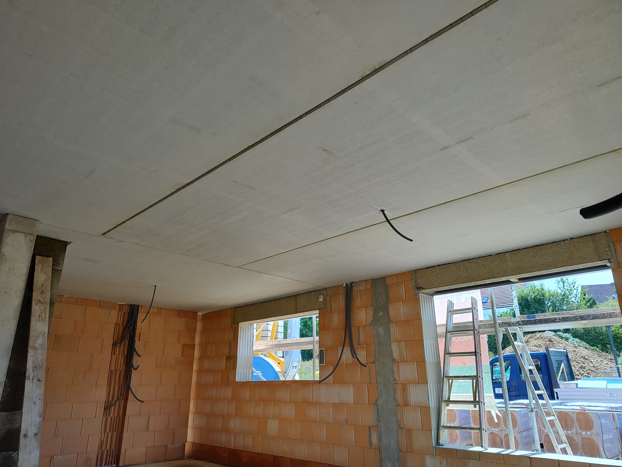 Surowy stan wnętrza domu w trakcie budowy: ściany z cegły, betonowy strop z widocznymi przewodami elektrycznymi, drabiny opierające się o okno.