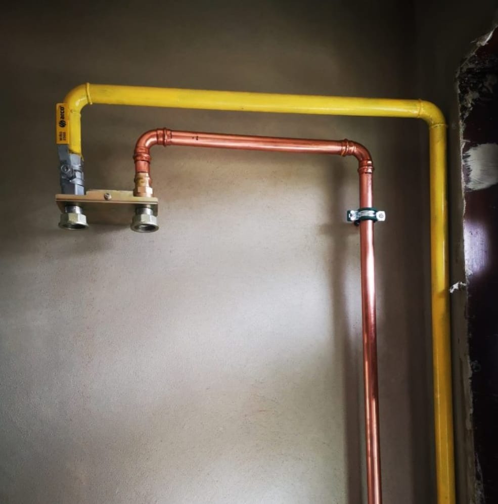 Żółta rura gazowa i miedziana instalacja wodna na szarej ścianie. Widoczne złącza i uchwyty mocujące. Instalacja połączona z zaworem.