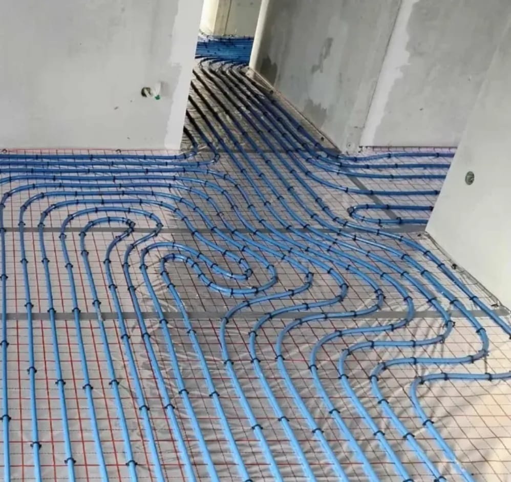 Niebieskie rury ogrzewania podłogowego ułożone na siatce w surowym wnętrzu. Widok z góry, ukazujący precyzyjny montaż instalacji przed wylaniem posadzki.