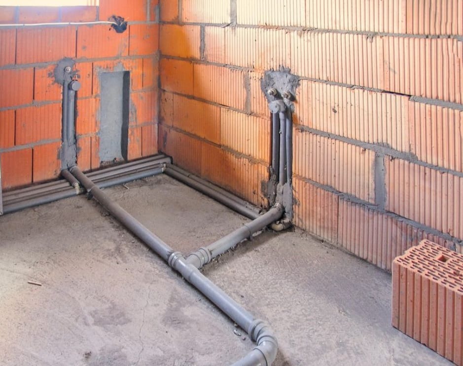 Szare rury instalacji hydraulicznej ułożone w surowym wnętrzu z ceglanymi ścianami. Widoczne łączenia i mocowania rur w ścianach. Etap prac przed zalaniem posadzki.