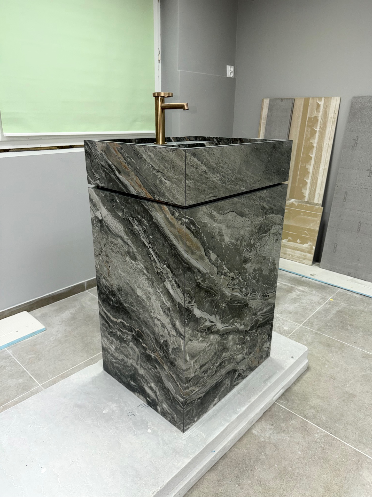 Stylowa, marmurowa umywalka kolumnowa z złotą armaturą w łazience w Olkuszu. W tle płytki i okno z zieloną roletą. Nowoczesny design.