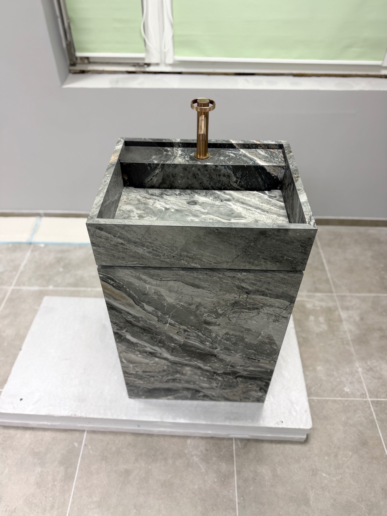 Nowoczesna, wolnostojąca umywalka z szarego marmuru z miedzianą baterią, ustawiona na białym podeście w łazience z szarą glazurą i oknem.