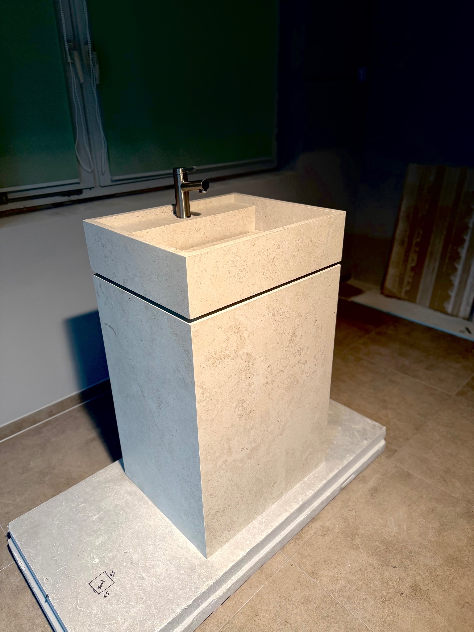 Beżowa, minimalistyczna umywalka wolnostojąca z chromowaną baterią, ustawiona na płytkach w surowym wnętrzu łazienki w trakcie wykończenia.