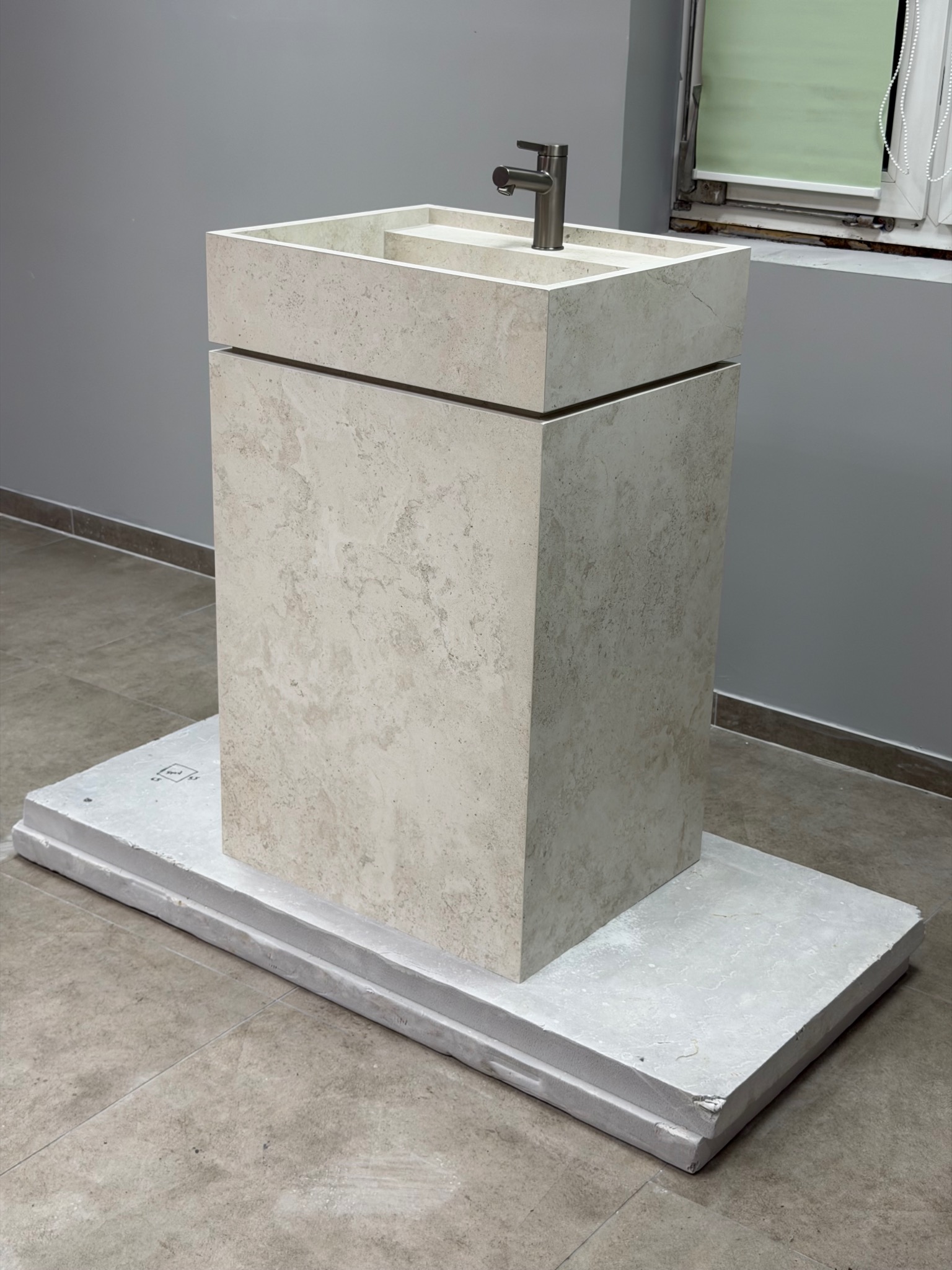 Nowoczesna, kamienna umywalka kolumnowa z chromowaną baterią, ustawiona na płycie. Szare ściany i okno w tle. Minimalistyczny design, surowy styl wykończenia.