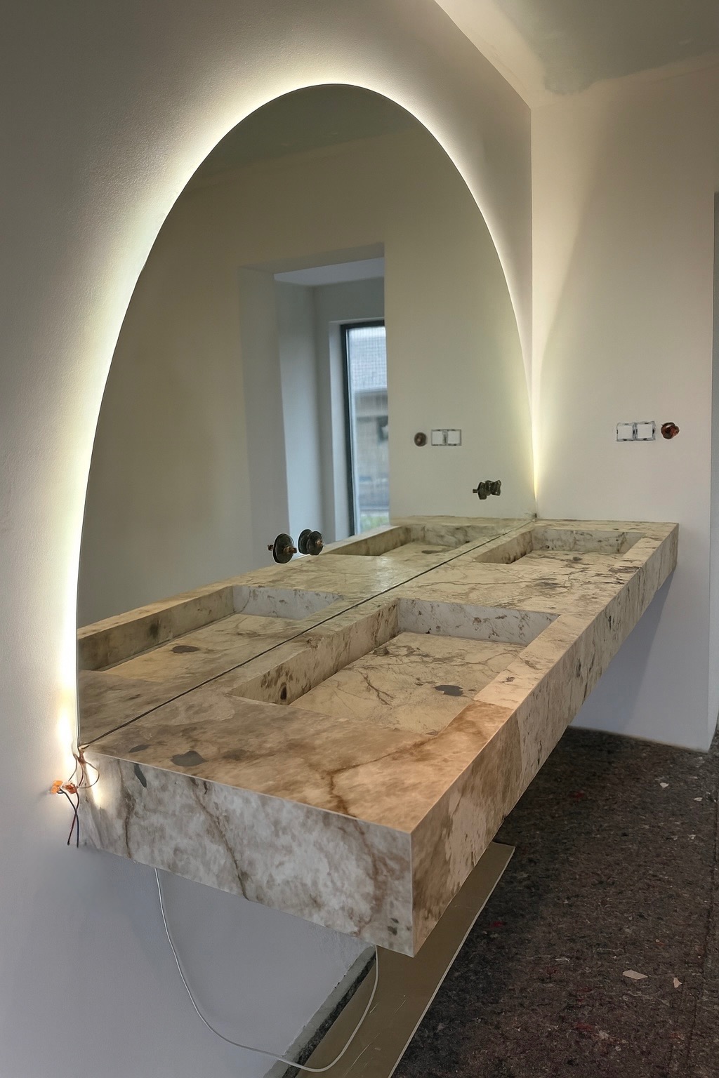 Nowoczesna łazienka z marmurową umywalką i podświetlanym lustrem w kształcie łuku. Widoczne niedokończone prace elektryczne.