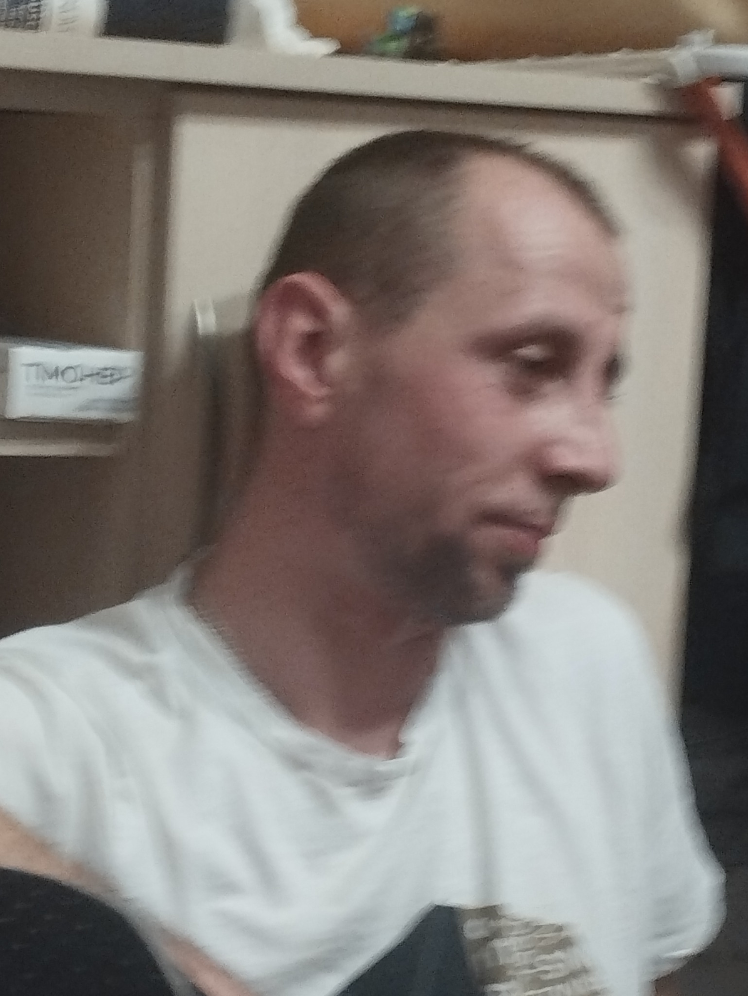 Portret mężczyzny z krótkimi włosami, ubranego w białą koszulkę, z profilu na rozmytym tle szafki i pudełka. Widoczny fragment łańcuszka na szyi.