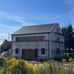 EU INVEST Daniel Krotochwil - Nowo otynkowany dom jednorodzinny w trakcie wykończenia, otoczony rusztowaniami i bujną roślinnością. Widoczne drewniane panele elewacyjne i ciemna dachówka.