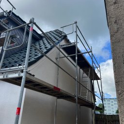 EU INVEST Daniel Krotochwil - Elewacja domu w trakcie prac wykończeniowych, widoczna z poziomu rusztowania. Świeżo otynkowana ściana, dachówka i fragment nieba z chmurami w tle.