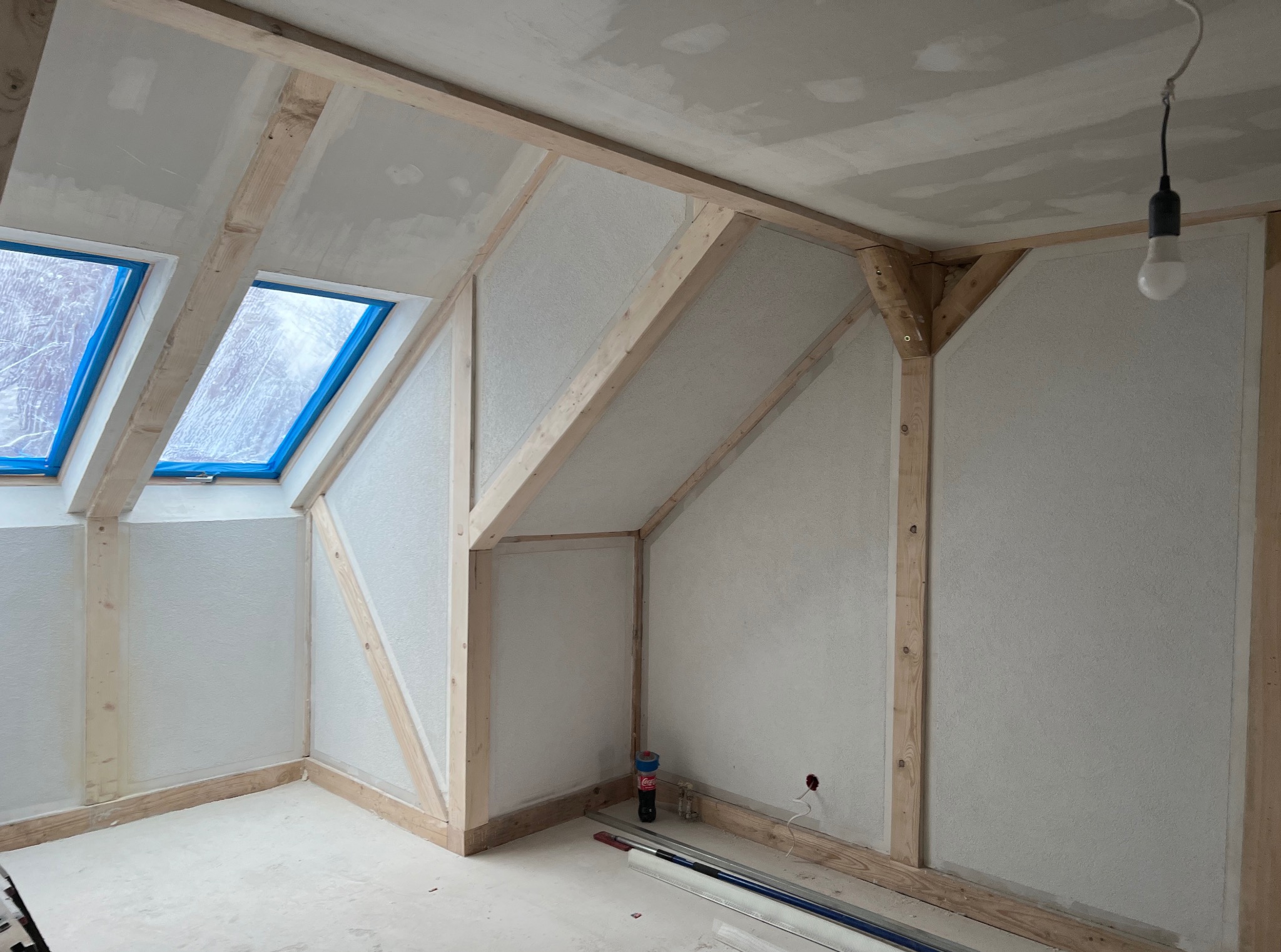 Poddasze w trakcie remontu: drewniana konstrukcja, okna dachowe zabezpieczone folią, ściany przygotowane do malowania, surowa żarówka. Widoczne materiały budowlane.