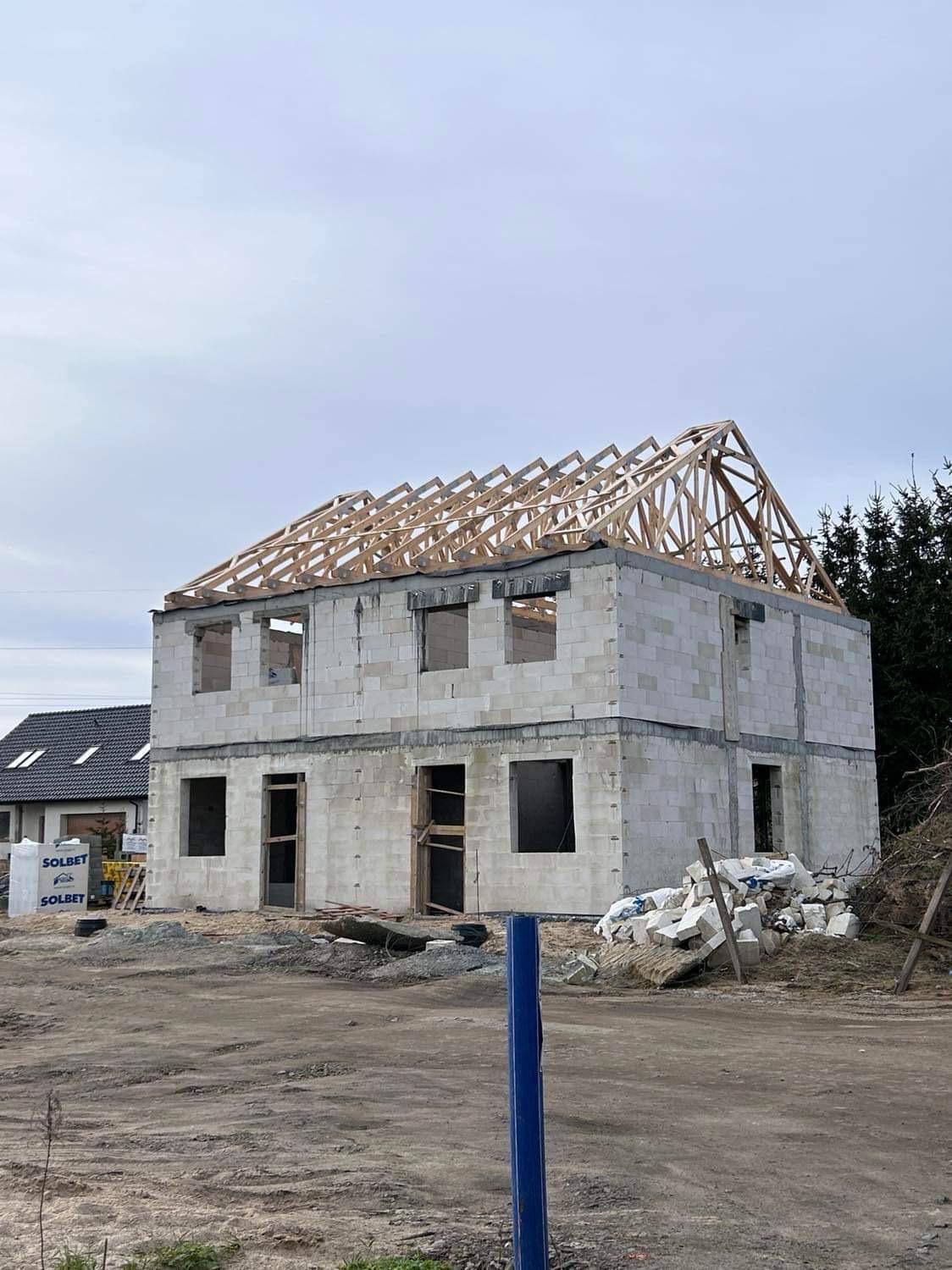 Surowy stan dwukondygnacyjnego domu z pustaków, z widoczną drewnianą konstrukcją dachu, worki z materiałami budowlanymi i gruz na placu budowy.