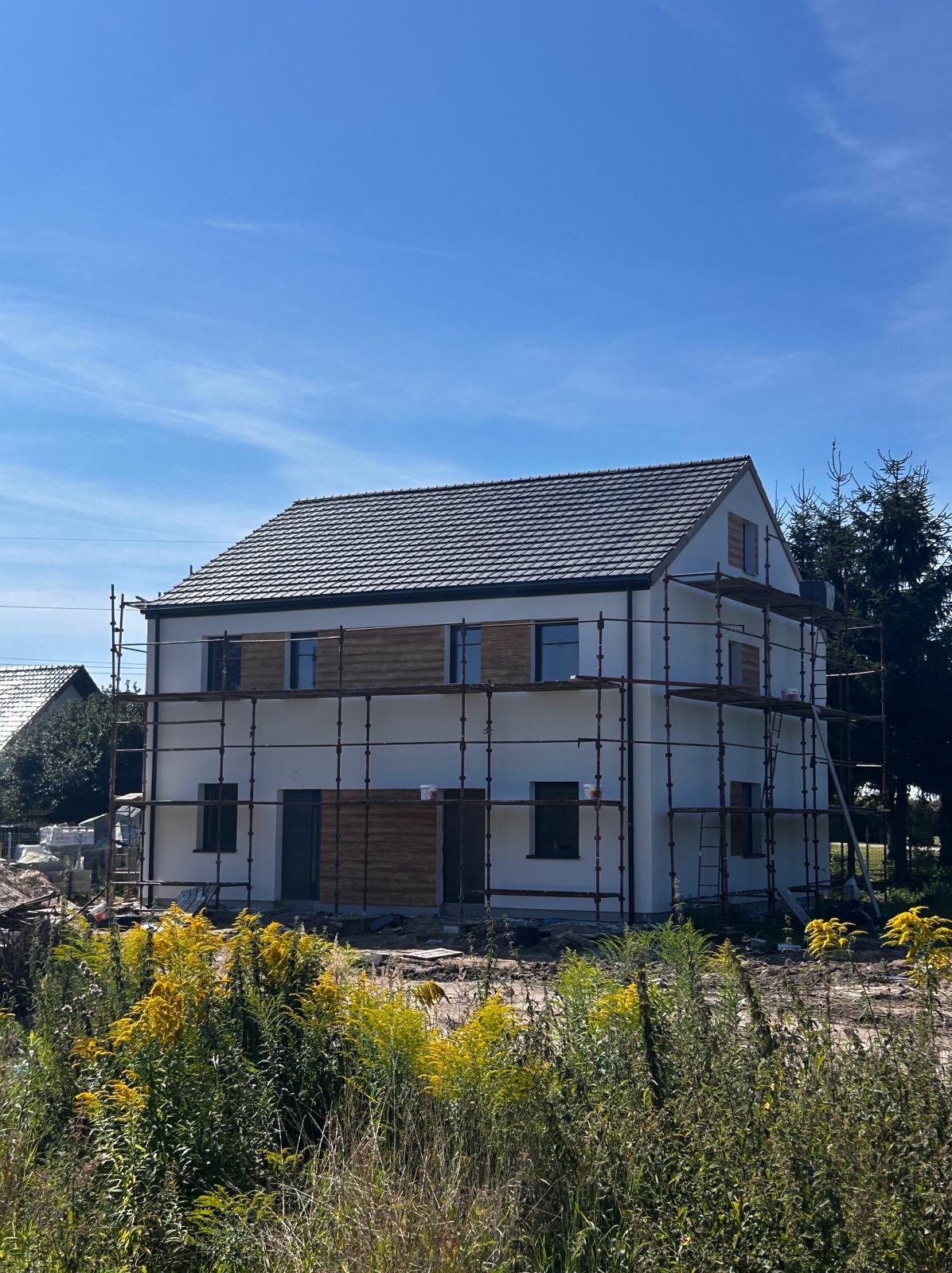 Nowo otynkowany dom jednorodzinny w trakcie wykończenia, otoczony rusztowaniami i bujną roślinnością. Widoczne drewniane panele elewacyjne i ciemna dachówka.