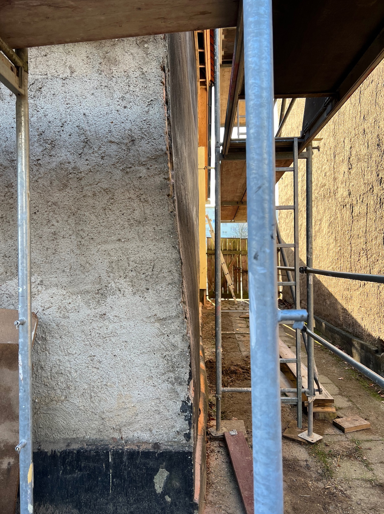 Ściana budynku w trakcie remontu, widoczna zza rusztowania. Szorstka faktura tynku, fragmenty czarnej farby u dołu. W tle drabina i ogrodzenie.