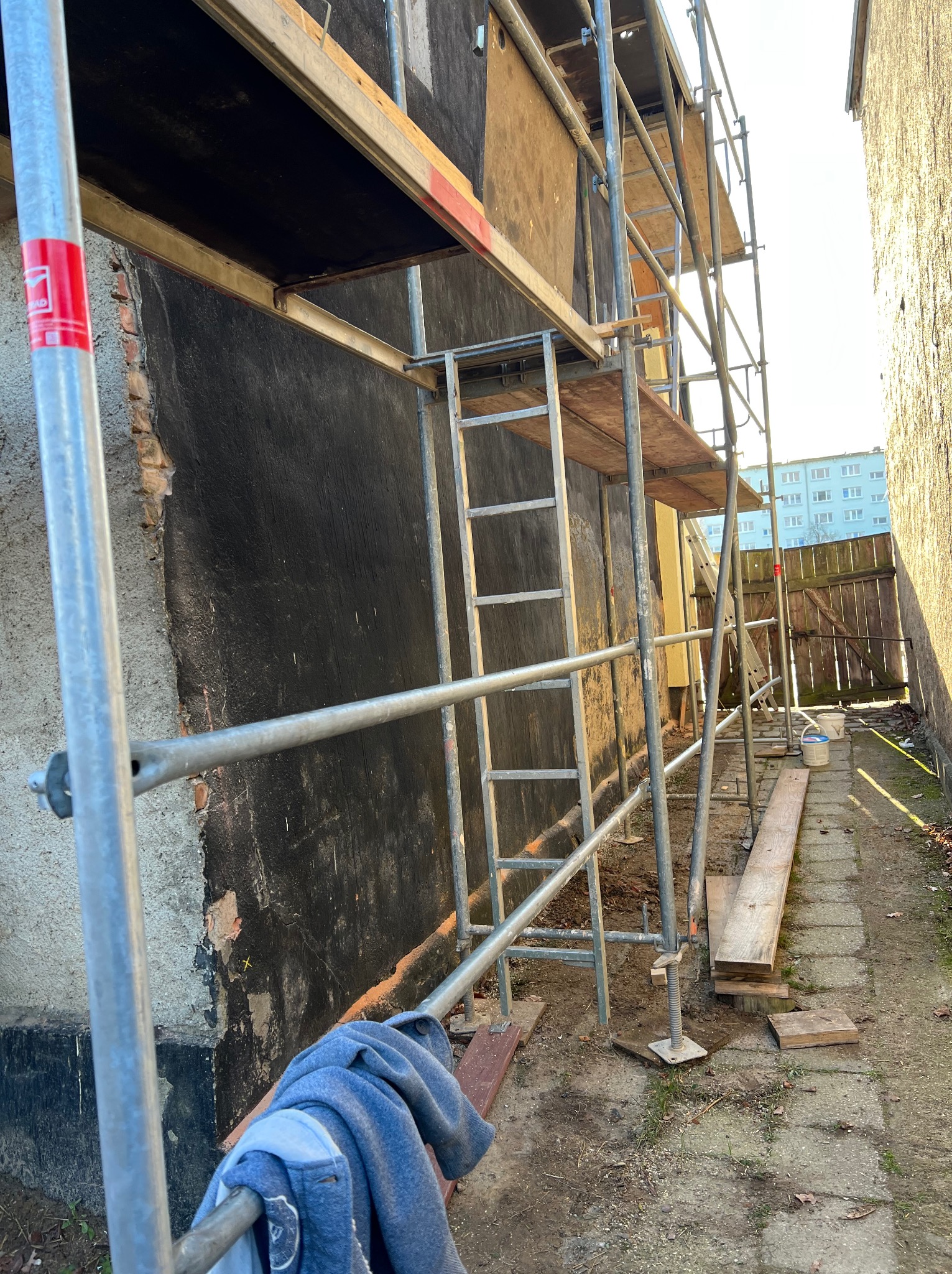 Ściana budynku w trakcie renowacji z rusztowaniem, widoczne ślady po usunięciu starej farby i przygotowanie do malowania. Na ziemi leżą deski i wiadro.