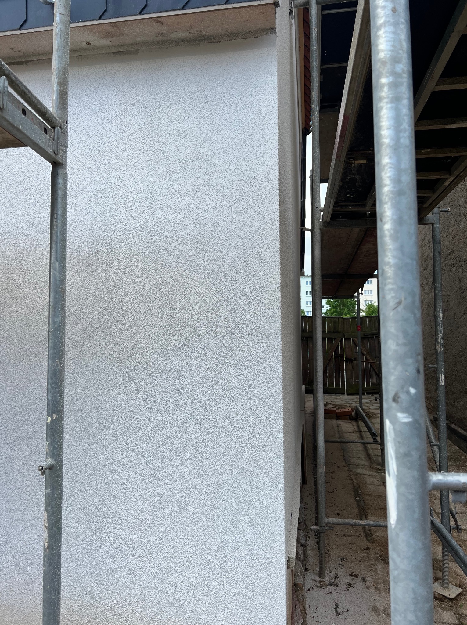 Świeżo otynkowana, biała ściana budynku z widocznym rusztowaniem. Tekstura tynku barankowa. Widok z dołu, fragment budynku w kadrze.