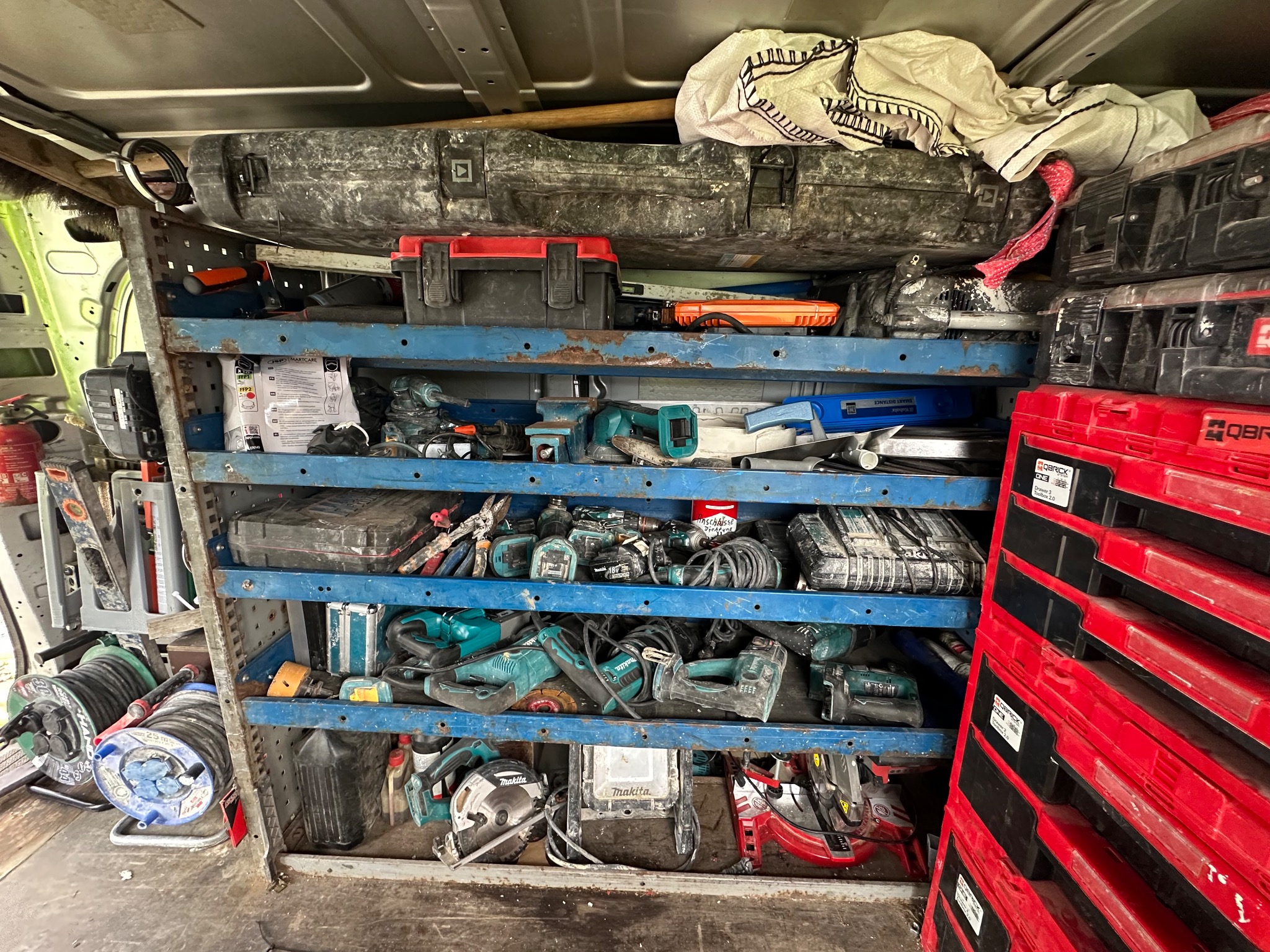 Półki w furgonetce wypełnione narzędziami i skrzynkami narzędziowymi, w tym wiertarki, piły i przedłużacze, z przewagą marki Makita. Widok z wnętrza pojazdu.