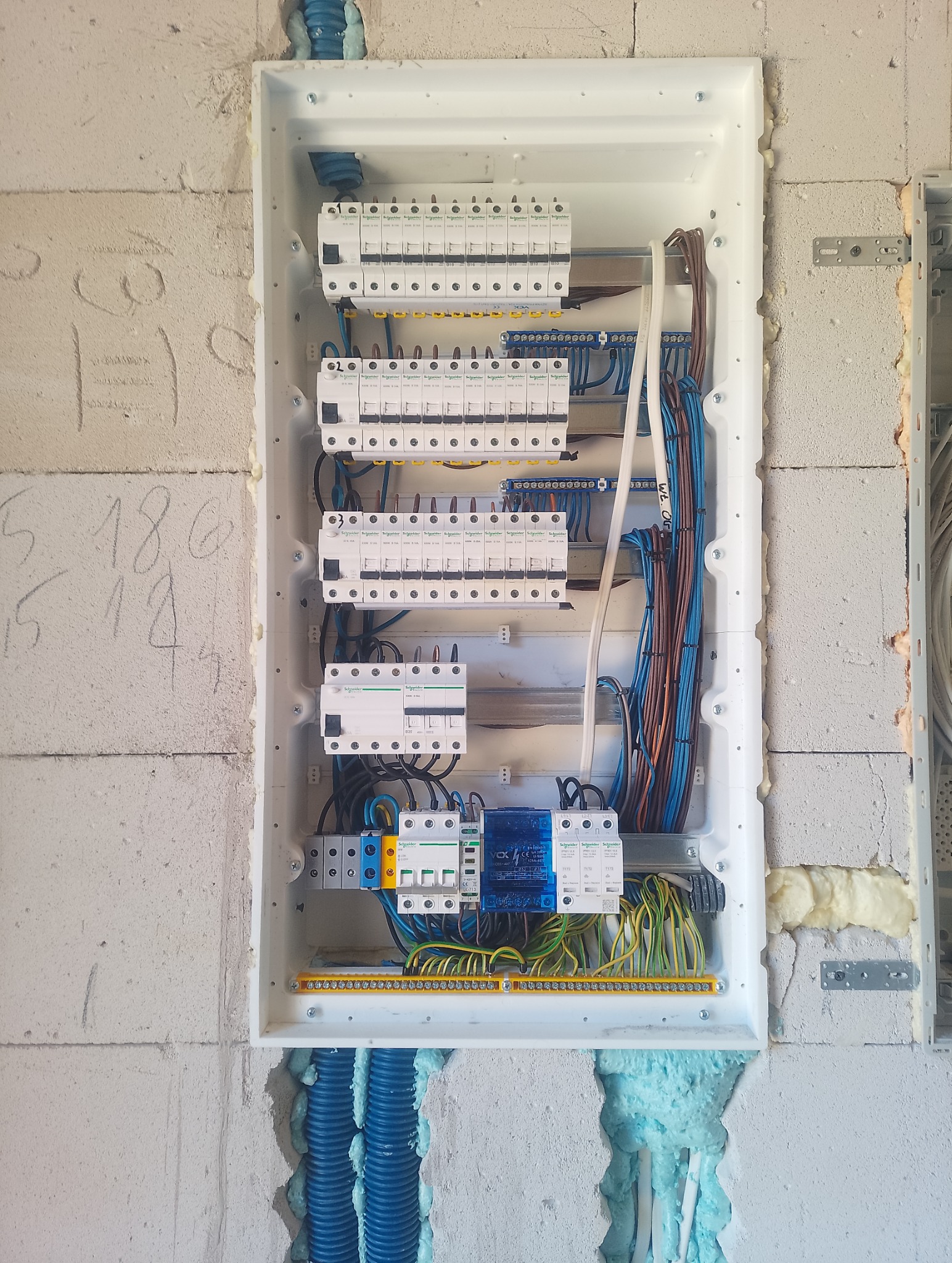 Rozdzielnica elektryczna w białej obudowie, zamontowana na surowej ścianie z betonu komórkowego, z widocznymi przewodami i zabezpieczeniami. Niebieskie rury instalacyjne.