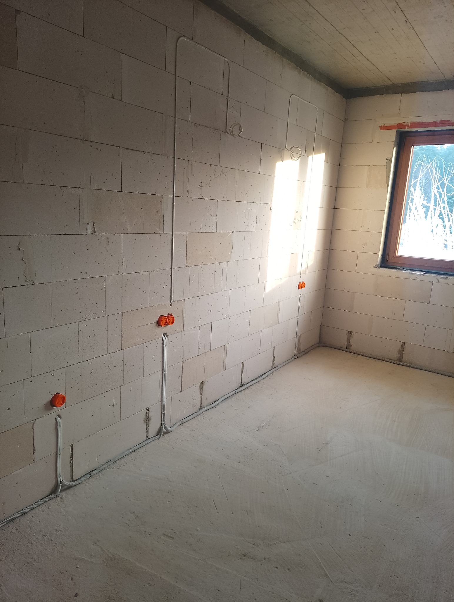 Surowy pokój z instalacją elektryczną w ścianach z betonu komórkowego. Widoczne peszle i puszki instalacyjne. W tle okno z widokiem na drzewa.