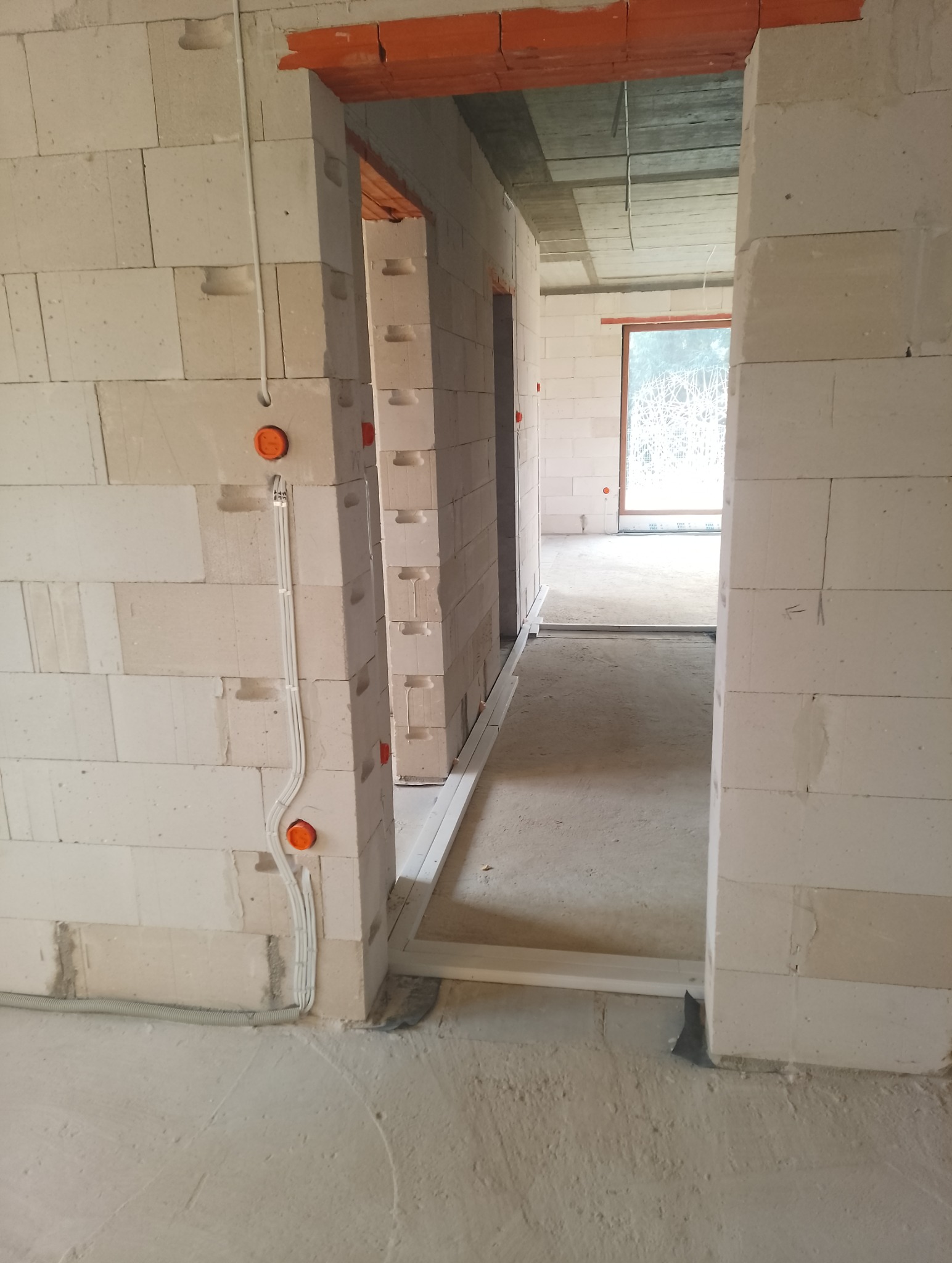 Wnętrze w stanie surowym: widoczne przewody elektryczne w ścianie z białych bloczków, przygotowane otwory na gniazdka. W tle widoczne okno i surowy betonowy strop.