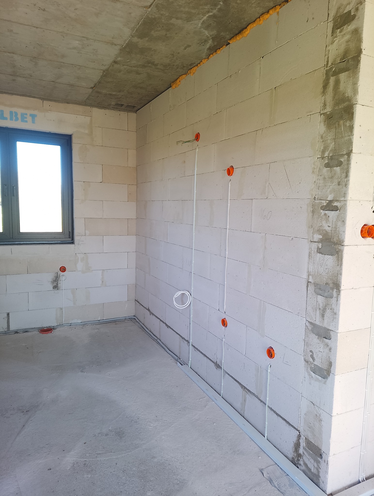 Surowy stan: instalacja elektryczna w nowym budynku z białych bloczków, widoczne kable w peszlach i puszki elektryczne. Okno po lewej stronie. Podłoga betonowa.