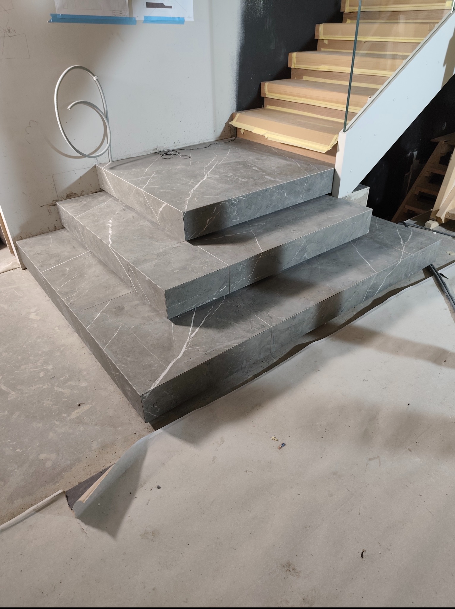Wykończone płytkami schody z szarym marmurkowym wzorem, widoczne w trakcie remontu. Częściowo zabezpieczone drewniane schody w tle.