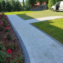 Ogrodowa Moc - Ułożona kostka brukowa tworząca ścieżkę w ogrodzie, obok rabata z różami i trawnik z ażurowymi płytami. W tle zaparkowany biały samochód dostawczy.