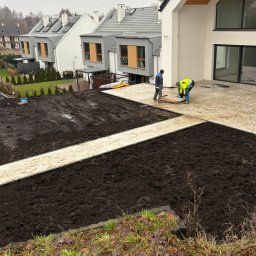 Ogrodowa Moc - Układanie ścieżki z płyt na przygotowanym gruncie pod trawnik, w tle nowoczesne domy. Prace ogrodowe w toku, widoczne narzędzia i dwóch pracowników.