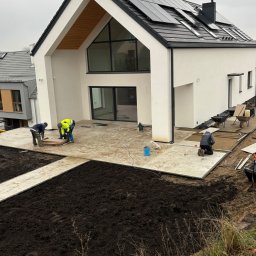 Ogrodowa Moc - Układanie kostki brukowej na posesji nowoczesnego domu w Świętajno. Pracownicy w trakcie realizacji ścieżki i tarasu. Widoczne narzędzia i materiały budowlane.