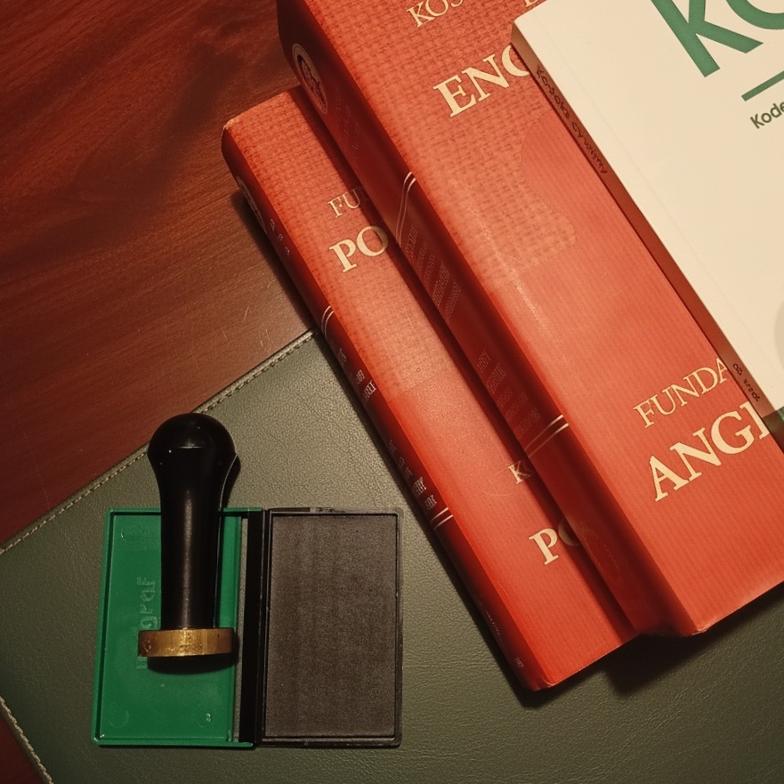 Stempel z tuszem obok książek z zakresu językoznawstwa, leżących na zielonej podkładce. Elementy biurowe tłumacza przysięgłego.