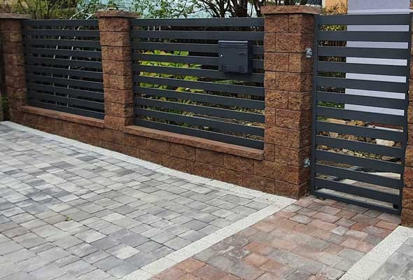 Nowoczesne ogrodzenie panelowe z ceglanymi słupkami i furtką, zintegrowane ze skrzynką na listy. Ułożona kostka brukowa w odcieniach szarości i brązu.