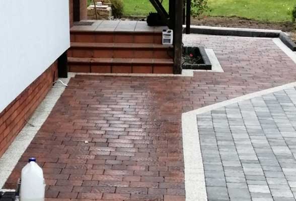 Ułożona kostka brukowa w dwóch odcieniach, brązowym i szarym, tworząca wzór na podjeździe. Widoczne schody wejściowe i fragment elewacji budynku. Kanister z płynem na kostce.