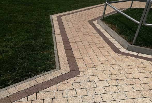 Ścieżka z beżowej kostki brukowej z ciemnobrązowymi akcentami, obrzeżona zielonym trawnikiem i elementami małej architektury. Widok z góry, równomierne oświetlenie.