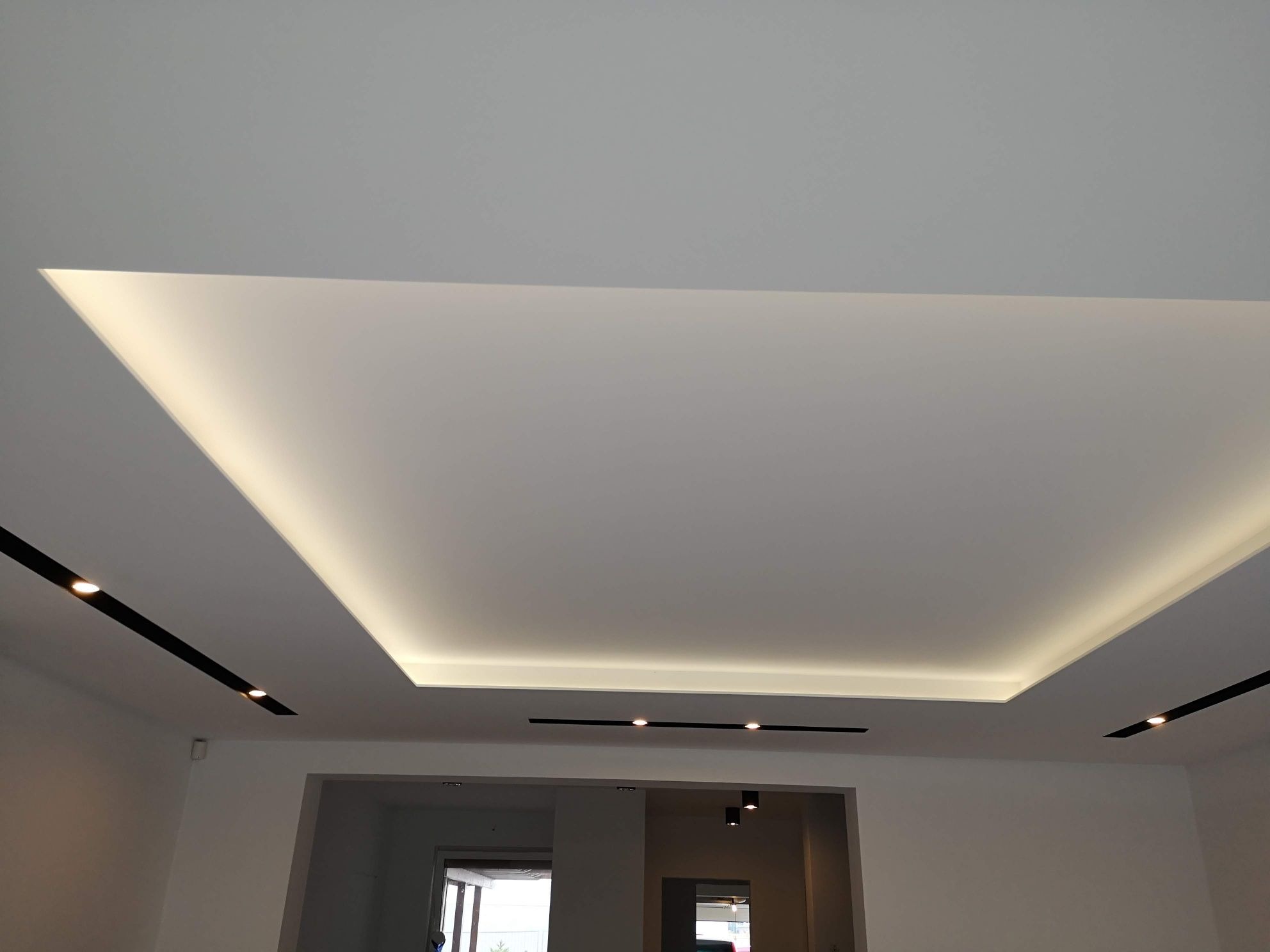 Nowoczesny sufit podwieszany z oświetleniem LED w kształcie prostokąta, czarne szyny z reflektorami, widok z dołu na białe ściany i przejście do innego pomieszczenia.
