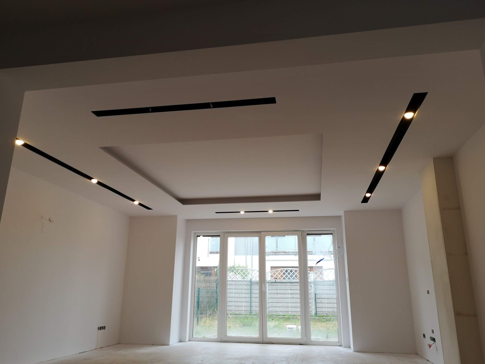 Nowoczesny sufit podwieszany z oświetleniem liniowym w przestronnym, jasnym pomieszczeniu z dużym oknem balkonowym. Minimalistyczny design.