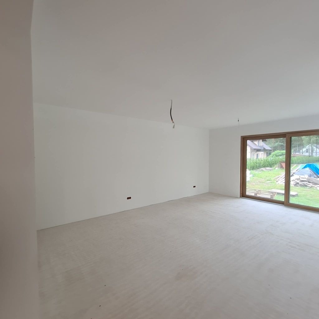 Jasne, przestronne wnętrze po szpachlowaniu ścian i wylewce podłogi, z widokiem na zewnątrz przez duże okno. Minimalistyczny design, surowy stan wykończenia.