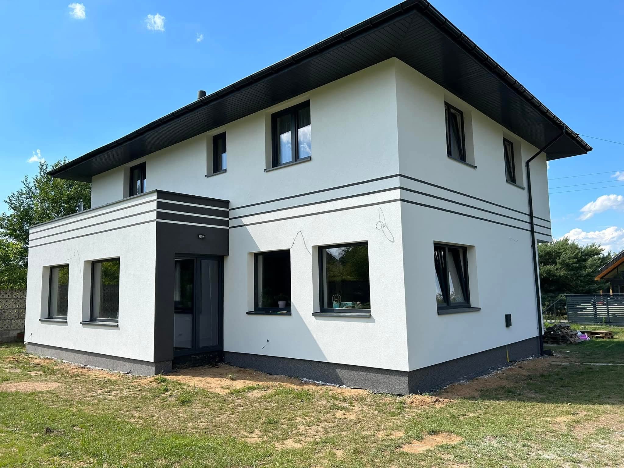 Nowoczesny dom z białą elewacją i czarnymi akcentami, widok z zewnątrz w słoneczny dzień. Minimalistyczny design z poziomymi pasami i ciemną stolarką okienną.