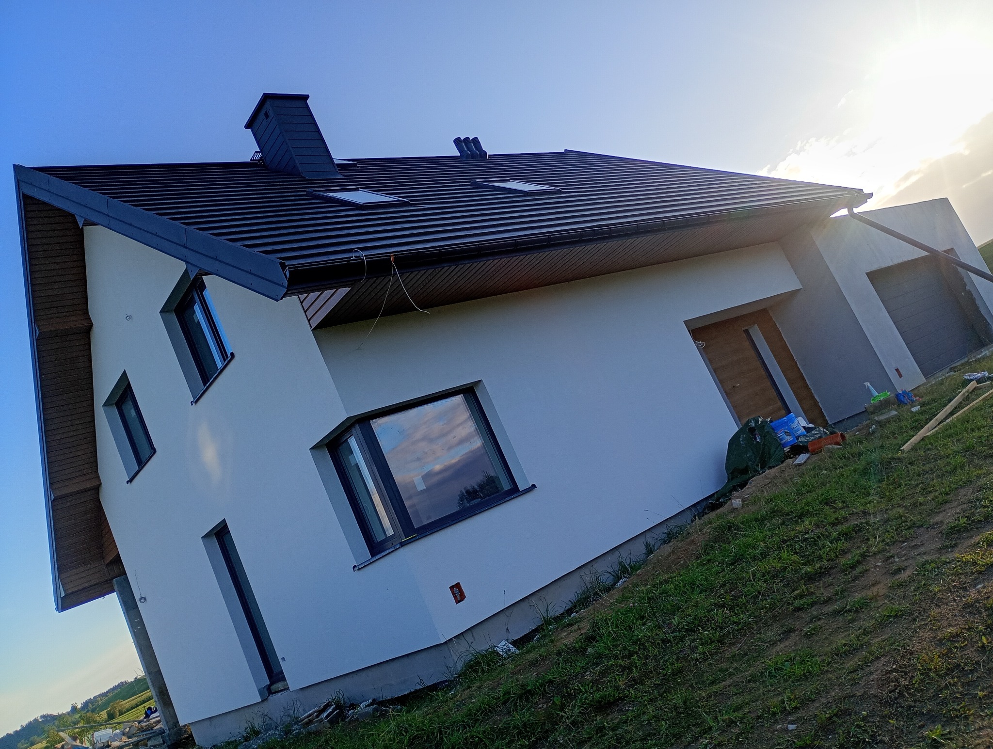 Nowoczesny dom jednorodzinny z ciemną dachówką i jasną elewacją w Szypliszkach. Widoczne okna z ciemnymi ramami i fragment ogrodu. Ukończony remont elewacji.